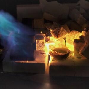 May include: A blue torch flame heats a small crucible with a metal object inside, surrounded by white ceramic pieces and glowing orange embers.
