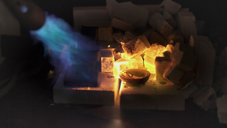 May include: A blue torch flame heats a small crucible with a metal object inside, surrounded by white ceramic pieces and glowing orange embers.