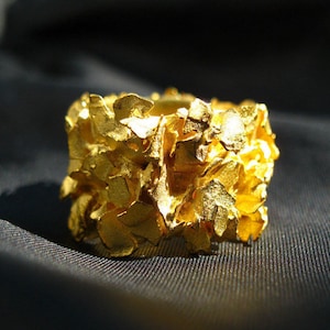 Op de afbeelding: Een gouden ring met een getextureerd, organisch ontwerp. De ring is gemaakt van veel kleine, onregelmatige stukjes goud die aan elkaar zijn gezet om een unieke en opvallende look te creëren.