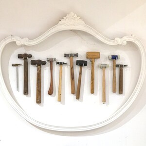 May include: A collection of 11 different hammers, including ball-peen hammers, rubber mallets, and a small metal hammer, are displayed on a white wall inside a decorative white frame.