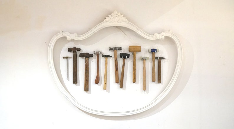 May include: A collection of 11 different hammers, including ball-peen hammers, rubber mallets, and a small metal hammer, are displayed on a white wall inside a decorative white frame.