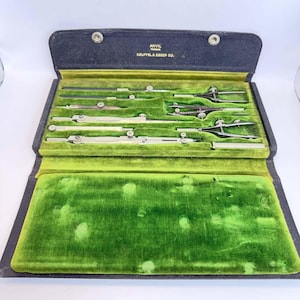 Può includere: Un set di strumenti da disegno tecnico in una custodia nera con interno in velluto verde. Il coperchio riporta la scritta ANVIL e KEUFFEL & ESSER CO. Il set include vari compassi, divisori e barre di prolunga in metallo per il disegno tecnico.