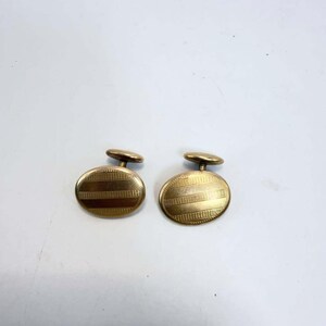 May include: A pair of oval-shaped gold-toned cufflinks featuring a horizontal striped pattern with alternating smooth and textured bands. Each cufflink has a central post and a small, rounded toggle mechanism on the back for secure fastening.