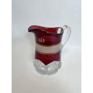 May include: A ruby red glass jug with a clear handle and base. The jug features a white band with gold trim and the number "1863" in white. The base has a cut glass design.