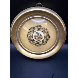 May include: A circular wall hanging featuring a gold-toned frame with a glass front. Inside, a central white porcelain medallion displays a painted scene of two figures, surrounded by an intricate, openwork gold-colored metal filigree border.