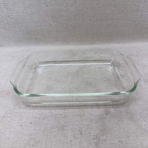 Puede incluir: Una fuente de horno rectangular de vidrio transparente con asas laterales integradas. El recipiente presenta una superficie lisa y transparente con un fondo plano, diseñado para su uso en el horno. El vidrio es incoloro y sencillo.