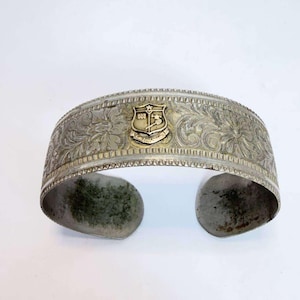 Puede incluir: Una pulsera brazalete ancha de tono plateado con un intrincado patrón floral en relieve en la banda. Centrado en la parte frontal hay un pequeño emblema dorado en forma de escudo con una estrella arriba y un gráfico de helicóptero.