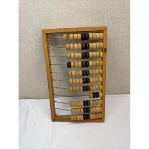 May include: A rectangular wooden abacus featuring ten horizontal metal rods. Each rod holds a series of round beads in natural light wood and dark brown tones, arranged to slide across the frame for counting and mathematical calculations.