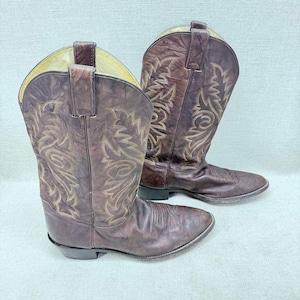 Op de afbeelding: Een paar donkerbruine leren cowboylaarzen met een spitse neus, een blokhak en ingewikkelde beige stikpatronen langs de schacht. De laarzen hebben treklipjes aan de bovenkant en een gladde, licht verweerde afwerking op het leer.
