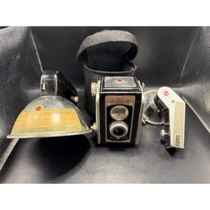 May include: A vintage Kodak Duaflex III camera with a black body and silver accents, accompanied by a flash unit, a light meter, and a black carrying case. The flash unit has a silver reflector and a yellowed label.