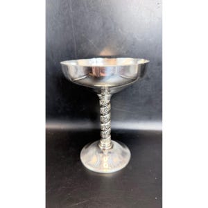 May include: A silver-colored champagne coupe with a wide, shallow bowl and a decorative, twisted stem. The base is round and the metal has a polished finish, reflecting the surrounding environment. The stem features an intricate design.