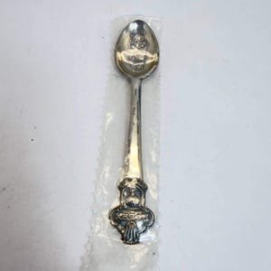 May include: A silver-toned metal spoon featuring an embossed Rolex crown logo and the text ROLEX on the handle. The bowl of the spoon displays a detailed engraved design. The item is encased in a clear, protective plastic sleeve for preservation.