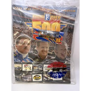 May include: A 2005 Food City 500 official program in a clear plastic sleeve, featuring a 1:64 scale die-cast replica race car in its original packaging, images of two race car drivers, and various racing-themed graphics and text throughout.