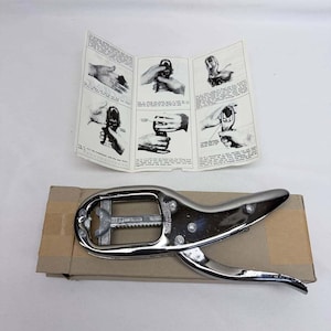 May include: A chrome-plated metal nutcracker tool with a ratcheting mechanism and a central bar marked with the year 1989. The tool is positioned on a rectangular cardboard box, accompanied by an instructional paper leaflet featuring six black and white diagrams.