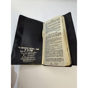 Peut inclure: Livre noir ouvert avec un dictionnaire et du texte de la Broadway National Bank of Bayonne, New Jersey. Le texte comprend l'adresse de la banque, le taux d'intérêt et l'adhésion à la FDIC.
