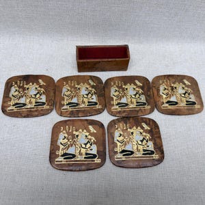 May include: A set of six square brown coasters with rounded corners and a matching rectangular storage box with a red interior. Each coaster features a gold and black Egyptian-style illustration of two figures and a bird, with a marbled wood grain finish.