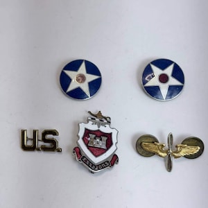 May include: A collection of five military-style pins including two circular blue and white star emblems, a gold-toned U.S. insignia, a shield-shaped crest with a red center and the text ESSAZONS, and a gold-toned winged propeller aviation pin.