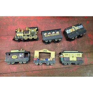 May include: A collection of miniature train cars, including a locomotive, passenger cars, and a caboose. The train cars are painted in shades of brown, yellow, and black, with details like windows and wheels. Some cars have the year "1998" on them.