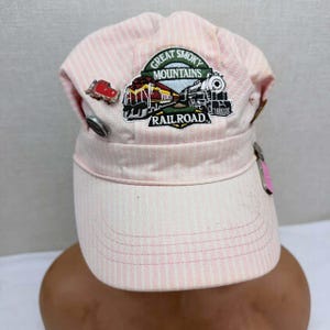 May include: A light pink and white pinstriped conductor hat featuring a central embroidered patch that reads Great Smoky Mountains Railroad with a train graphic. The hat is adorned with several metal pins, including a red train engine pin.