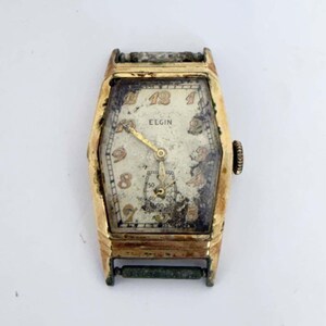 May include: A vintage gold-tone Elgin wristwatch with a rectangular case and a weathered, off-white dial. The face features gold-colored Arabic numerals, a sub-dial for seconds at the bottom, and gold hands. The metal shows significant patina.