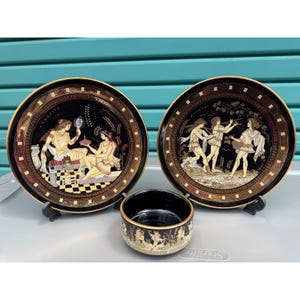 May include: A set of three black decorative ceramic pieces with gold trim. The set includes two plates and a small bowl. The plates feature classical Greek figures and text. The bowl has a similar design. The plates are displayed on black stands.