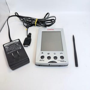 May include: A silver Compaq Aero personal digital assistant with a grey screen, four circular buttons, and the text "Powered by Microsoft Windows CE." Included are a black power adapter with a long cord and a thin black stylus pen for navigation.