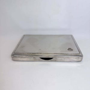 May include: A rectangular silver-toned metal case with a smooth, polished finish and a beveled edge. The top surface features a small, engraved geometric logo consisting of three interlocking triangles in the bottom right corner and a central notch.