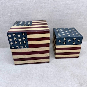 Pode incluir: Duas caixas quadradas de madeira com um design de bandeira americana desgastada nas cores vermelho, branco e azul. A caixa maior está à esquerda e a menor à direita, ambas exibindo um campo azul com estrelas e listras horizontais.