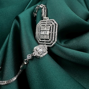 Könnte beinhalten: Ein silbernes Halsketten- und Armband-Set mit geometrischem Design. Der Anhänger und das Armband weisen rechteckige und quadratische Formen auf, die mit kleinen, funkelnden Steinen besetzt sind. Der Schmuck wird auf einem dunkelgrünen Satin-Stoff präsentiert.