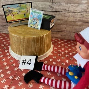 May include: A miniature scene featuring a small TV, a video game case, and a black box labeled "Box". The scene is set on a wooden platform with a red and white snowflake patterned background. The number "#4" is on a white card.