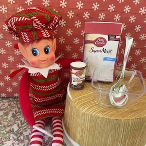 Puede incluir: Una muñeca elfo navideña con un atuendo y sombrero a rayas rojas y blancas, con zapatos negros. Junto a la muñeca hay suministros para hornear: una caja de mezcla para pastel blanco, un pequeño recipiente de glaseado Rich & Creamy, un cuenco transparente y una cuchara.