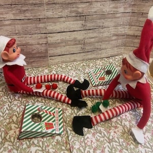 May include: Two elf dolls in red and white striped outfits with black shoes and white collars are playing a miniature Elf Corn Hole Game. The game includes small green and white striped boxes with a hole for tossing bean bags. Red pom-poms are scattered around.