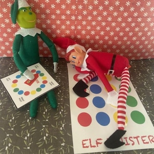 May include: Two elf dolls playing a game of Twister. One green elf holds the game board, the other red elf lies on the mat. The mat has coloured circles and the words "ELF SISTER". A festive scene with a red snowflake background.