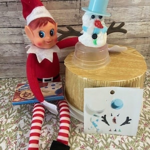 May include: A red and white elf doll with a matching hat and striped legs sits beside a small snowman figure. The snowman has a blue top hat and twig arms. A box of cookies and a craft sheet are also present.