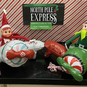 May include: Two elf dolls sitting in inflatable toy airplanes. One elf wears red, the other green. The planes feature candy cane patterns and propellers. A sign above reads North Pole Express Arrivals Just In Time For Christmas.