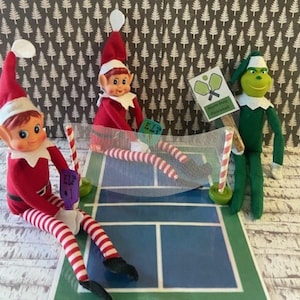 May include: Three Christmas elf dolls playing pickleball on a miniature court. The elves are wearing red and white outfits, and one is dressed as the Grinch. A sign reads "North Pole Pickleball".