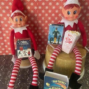 May include: Two elf dolls wearing red and white outfits, red hats, and black boots. One holds a "Charlie Brown Christmas" book, the other holds a "Elf" movie and a popcorn container. A box of "Pop Secret" popcorn is also visible.