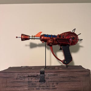 May include: A red and blue ray gun prop with a black handle, displayed on a wooden box. The ray gun has a futuristic design with a long barrel, fins, and a circular component. The wooden box has text and a metal clasp.