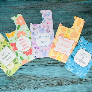 May include: Five colorful phone stands with floral patterns and inspirational quotes. The stands feature phrases like "Give Yourself Grace," "Choose Joy," and "Attitude of Gratitude." The background is a distressed teal wood surface.
