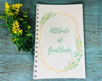 May include: A spiral-bound notebook with the words "Attitude of Gratitude" in a script font, surrounded by a gold geometric frame and watercolor leaves. Yellow and green floral sprigs are on the left side, set against a teal wooden surface.