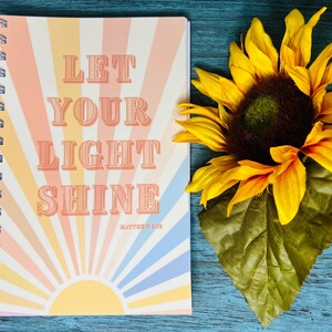 May include: A spiral-bound notebook with the words "LET YOUR LIGHT SHINE" in a retro font, with a sunburst design in pastel colors. A large sunflower with yellow petals and a green leaf is next to the notebook.