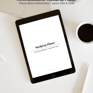 Könnte beinhalten: Ein schwarzes Tablet mit dem Text "Mealprep Planer" und "Plane deine Mahlzeiten - spare Zeit & Geld" auf weißem Hintergrund. Ein weißer Stift, eine Tasse Kaffee und ein Laptop liegen auf einer weißen Oberfläche.