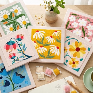 Peut inclure: Plusieurs broderies florales colorées. Chaque toile carrée présente un motif floral différent, notamment des marguerites et d'autres fleurs, dans des tons de jaune, rose, bleu et vert. Du fil à broder et une aiguille à punch sont également visibles.