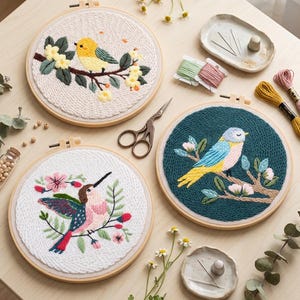 May include: Three embroidered bird designs in wooden hoops. One features a yellow bird on a branch with yellow flowers, another a blue and yellow bird, and the third a pink and blue hummingbird. Embroidery supplies are also visible.