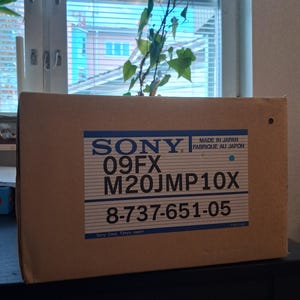 May include: A rectangular brown cardboard box featuring a white label with blue horizontal lines and text that reads SONY, 09FX, M20JMP10X, 8-737-651-05, MADE IN JAPAN, FABRIQUE AU JAPON, and Sony Corp. Tokyo Japan, with a small blue dot on the right side.