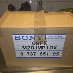 May include: A cardboard box with a blue and white label that reads "SONY" and "MADE IN JAPAN FABRIQUE AU JAPON". The label also displays the codes "09FX", "M20JMP10X", and "8-737-651-05". The box is sealed with clear tape.