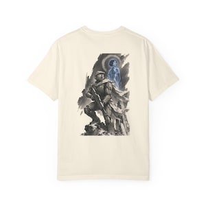 Könnte beinhalten: Cremefarbenes T-Shirt mit einer detaillierten Illustration eines Soldaten in Rüstung, der ein Gewehr hält, mit einer geisterhaften Figur im Hintergrund. Das Kunstwerk ist in Grau- und Blautönen gehalten, vor einem dunklen Hintergrund.