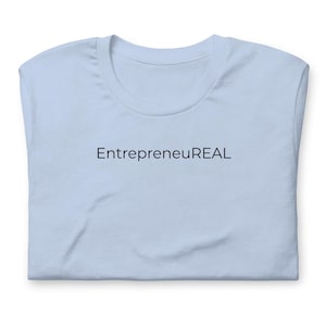 May include: A light blue t-shirt with the word "EntrepreneuREAL" printed in black across the chest. The shirt is folded, showing the neckline and sleeves.