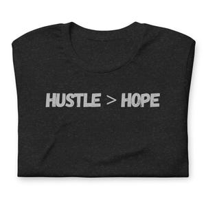 May include: Dark gray t-shirt with the text "HUSTLE > HOPE" printed in white. The shirt is folded and has a crew neck. The text is in a bold, sans-serif font.