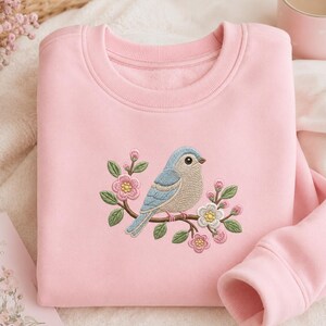 Può includere: Una felpa rosa chiaro con un ricamo di un uccello azzurro appollaiato su un ramo fiorito. L'uccello ha ali azzurre e corpo beige. I fiori sono rosa e bianchi con foglie verdi. Una tazza rosa è sullo sfondo.